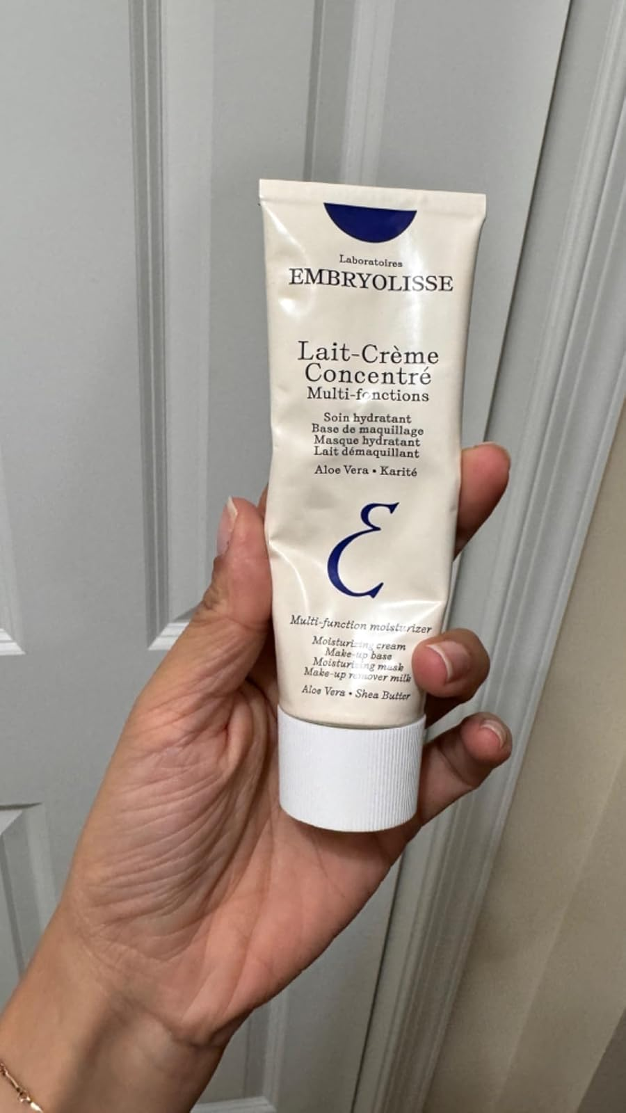 Hand holding a conduit  of Embryolisse Lait-Crème Concentré, a multi-function moisturizer with aloe vera and shea butter, against a airy  background