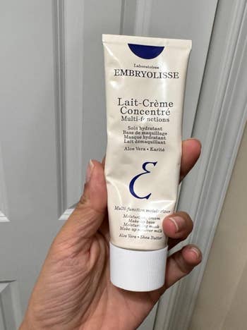 Hand holding a conduit of Embryolisse Lait-Crème Concentré, a multi-function moisturizer with aloe vera and shea butter, against a airy background