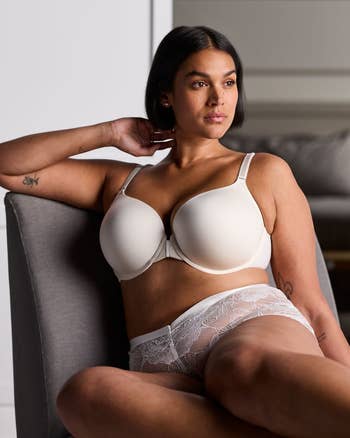 Woman in a beige bra and lace underwear seated on a sofa, posing for a lingerie ad