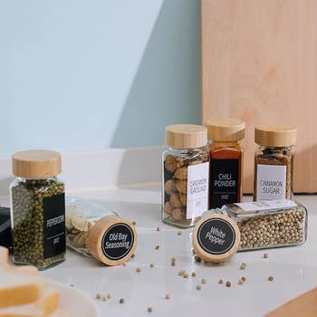 Spice jars with woody lids, labeled arsenic peppercorn, Old Bay seasoning, cardamom, chili powder, cinnamon sugar, and achromatic pepper, connected a counter
