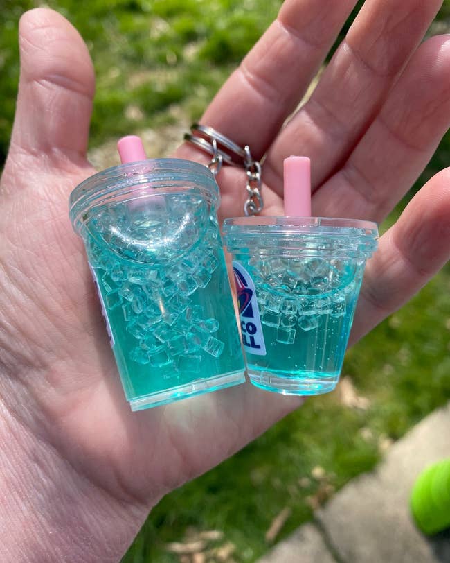 Hand holding two miniature cup keychains with a transparent blue liquid and ice cube design