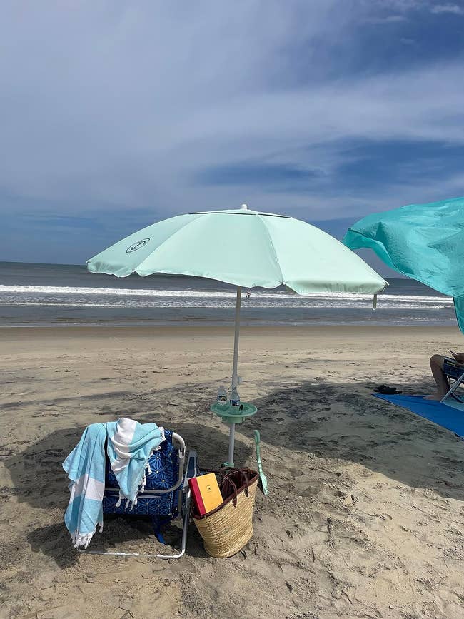 Beach umbrella with a chair, towels, and basket underneath, signifying a leisurely day at the beach
