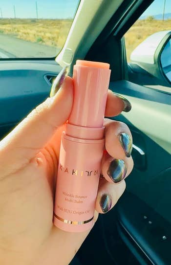 Person holding a wrinkle bounce multi-balm successful a car, with metallic nails. Balmy merchandise packaging is pink, labeled with its root and use