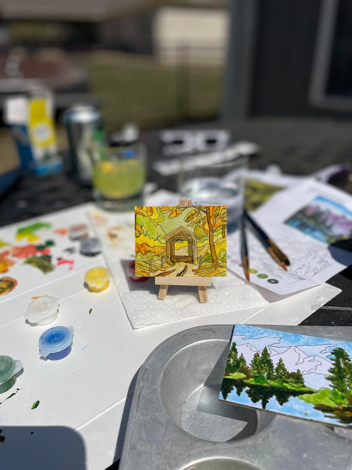 Mini painting of a fall scene on a small easel, surrounded by art supplies on a table outdoors