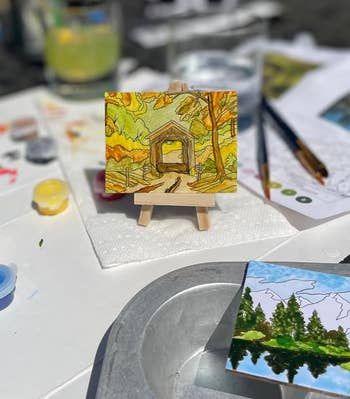 Mini painting of a fall scene on a small easel, surrounded by art supplies on a table outdoors