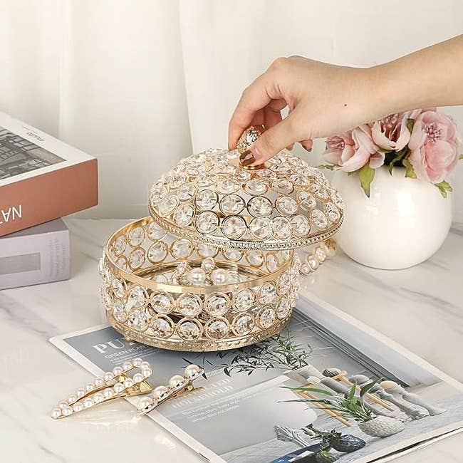 model opens a bejeweled round storage box on a marble table with accessories