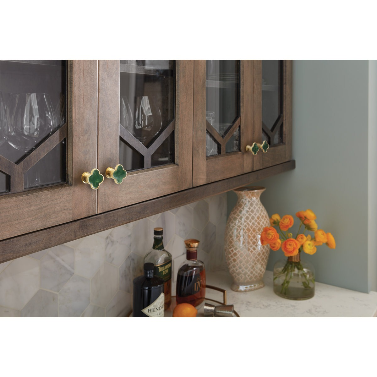 Kitchen cabinet with wooden glass-paneled doors, decorative green knobs, a patterned vase with orange flowers, and bottles on a marble countertop