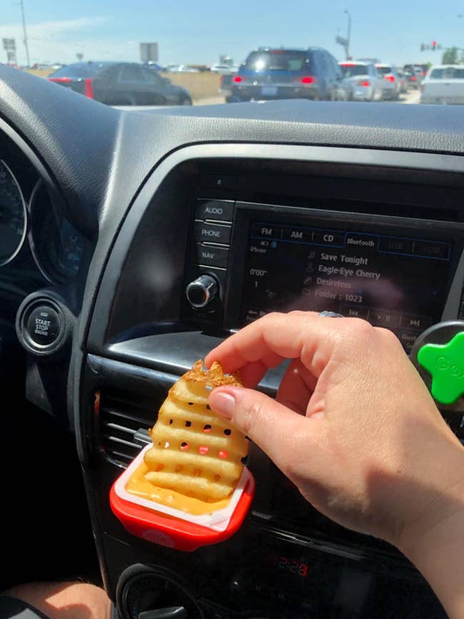 reviewer photo of them dipping a waffle fry into sauce held by the clip-on saucemoto attached to their car vent