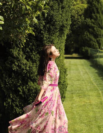 model in a floral dress with a pink belt, standing by greenery, looking upwards