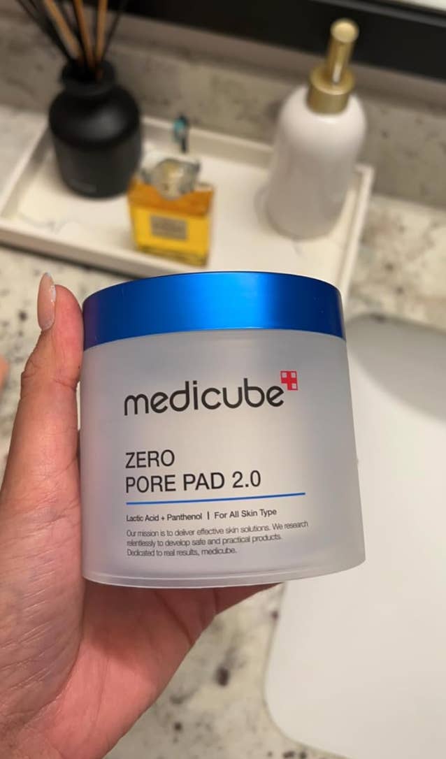 Hand holding Medicube Zero Pore Pad 2.0 container, a skincare merchandise  for treating pores. Background includes bath  items