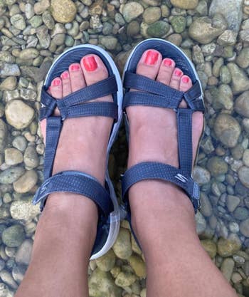 reviewer wearing the sandals in dark gray on a pebbly surface