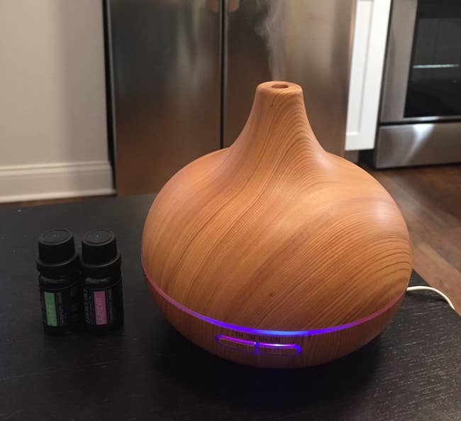 reviewer photo of diffuser next to some essential oils, emitting steam and lighting up purple