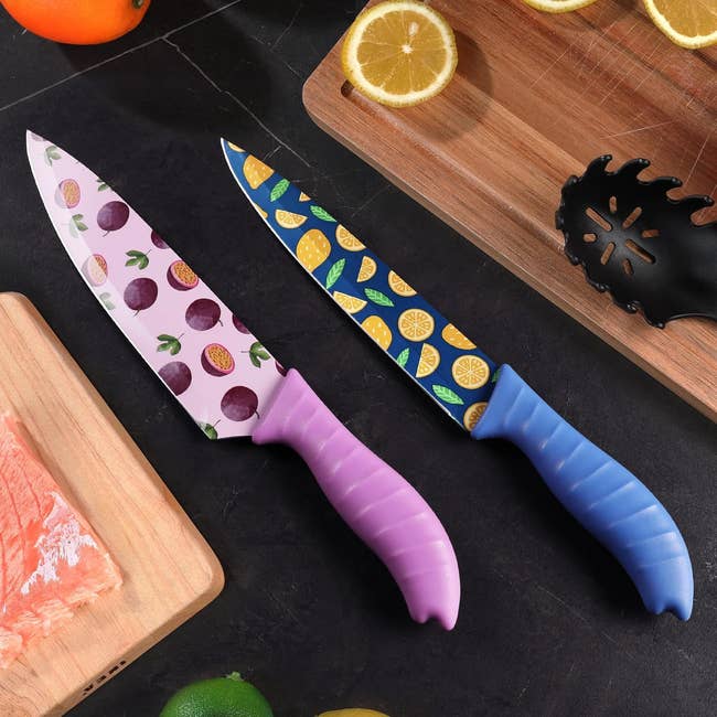 Two room knives with fruit-patterned blades, 1 with a pinkish grip and the different with a bluish handle, placed connected a countertop adjacent to a cutting committee with a citrus and salmon