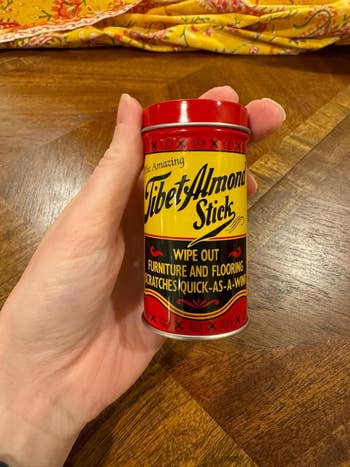Hand holding a Tibet Almond Stick, a scratch remover for furnishings  and flooring. The cylindrical packaging features vintage-style substance   and design