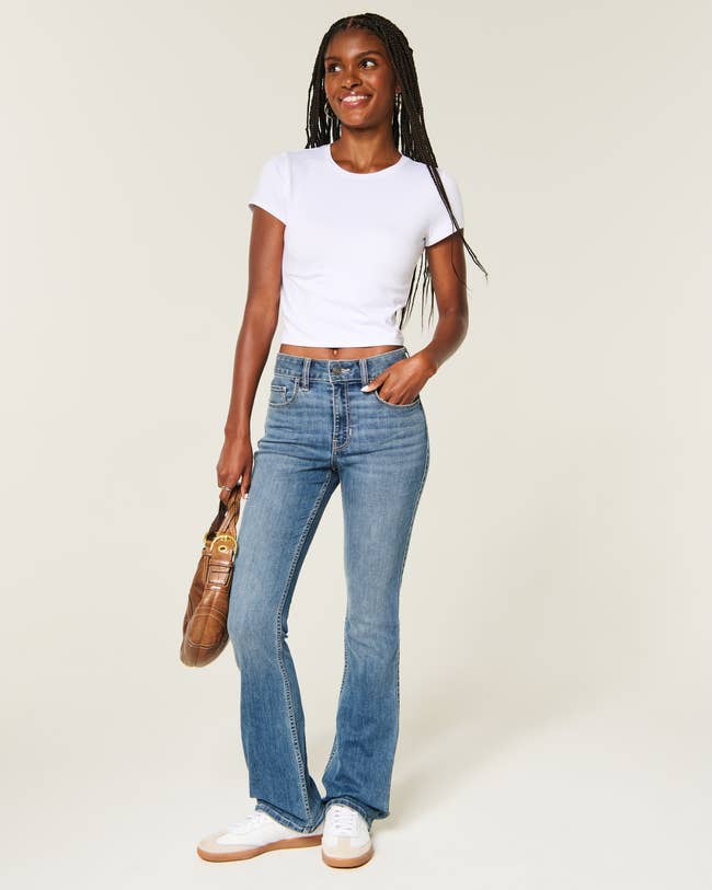 Person successful casual outfit with a achromatic tee, bootcut jeans, sneakers, and a brownish handbag, smiling and lasting against a neutral background