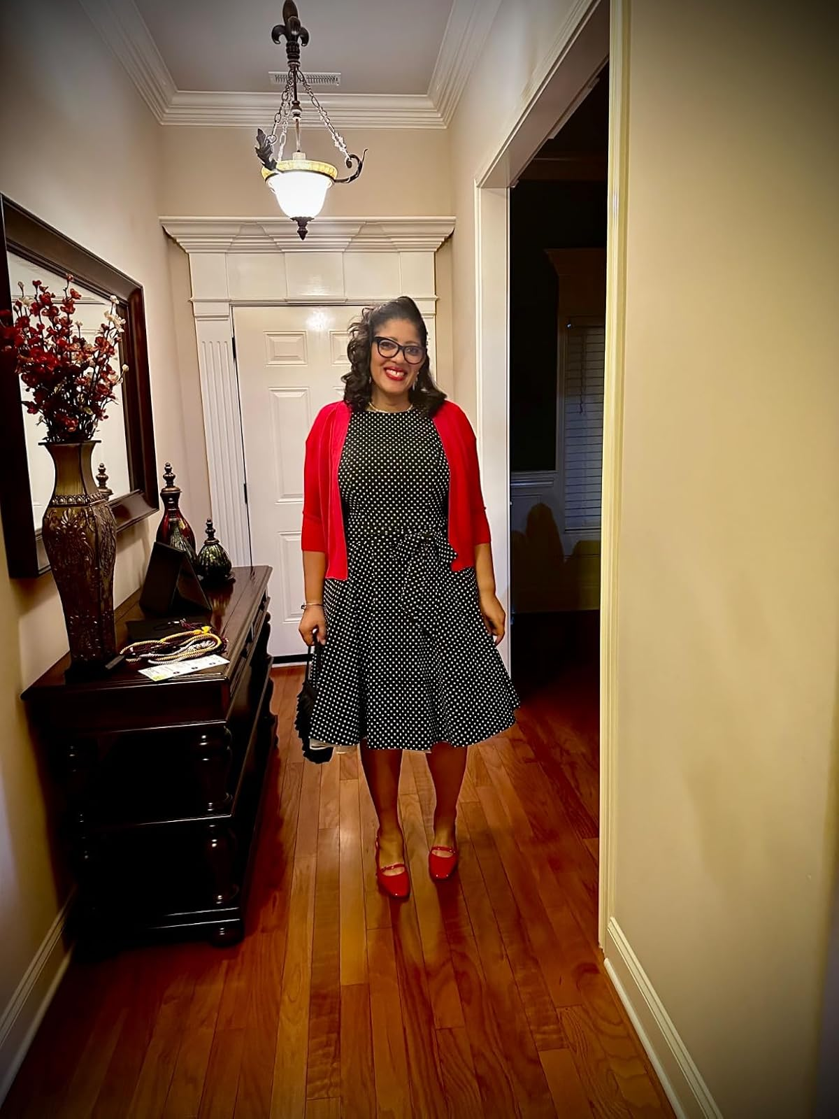 Person in a black and white polka dot dress with a red cardigan and red shoes stands in a hallway, smiling, holding a small black handbag