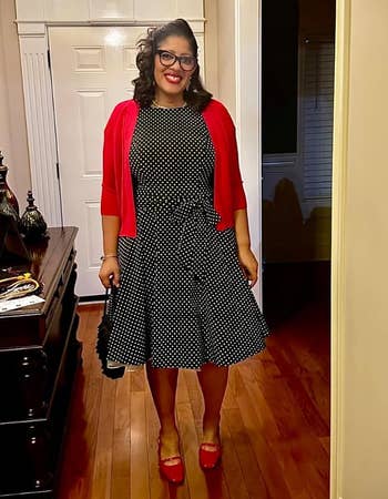 Person in a black and white polka dot dress with a red cardigan and red shoes stands in a hallway, smiling, holding a small black handbag