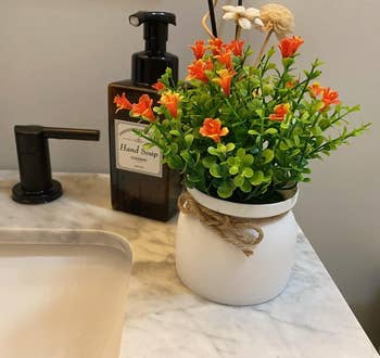 Artificial orange flowers in a white pot with a rustic bow, next to a sink