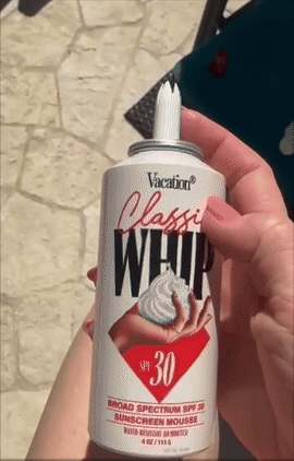 Person holding a can of Vacation Classic Whip SPF 30 sunscreen mousse, resembling whipped cream, with a poolside background
