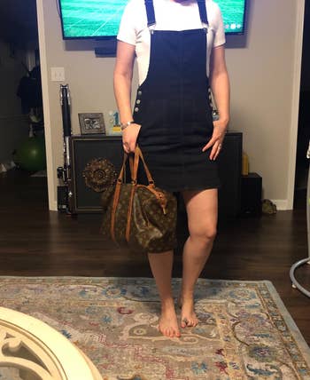 Person wearing a black denim overall dress with a white shirt, holding a designer handbag in a living room setting