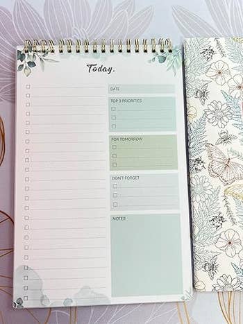 A spiral-bound daily planner with sections for date, priorities, tomorrow's tasks, reminders, and notes, on a floral background