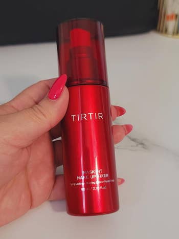 Hand holding a reddish  vessel  of Tirtir Mask Fit Makeup Fixer, a long-lasting mounting  spray for makeup