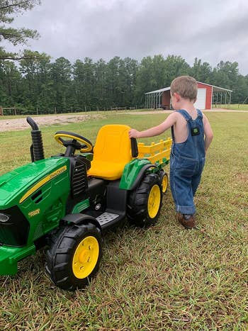 Child in overalls standing next to a toy John Deere tractor in a grassy field