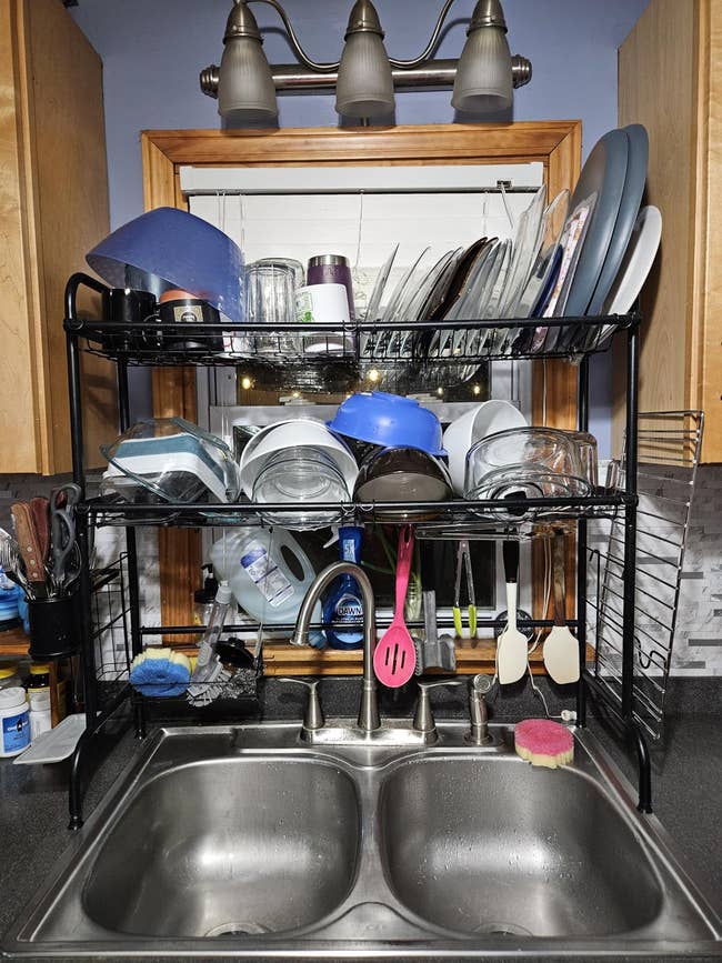 Kitchen sink with double-basin, drying rack loaded with dishes and utensils, overhead light fixture, and various cleaning supplies on counter