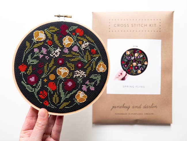 Cross-stitch kit featuring floral embroidery signifier with a manus holding the finished piece, labeled