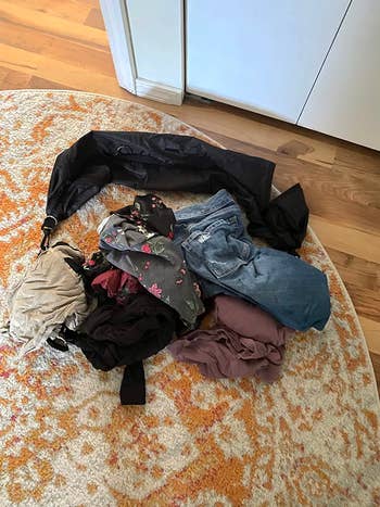 A pile of clothes on the floor next to a black unzipped tube 