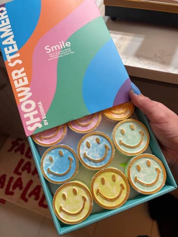 reviewer holding an open box of Shower Steamer by Smile, featuring scented tablets with smiley face designs