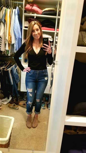 A reviewer taking a selfie in a closet, wearing a black long-sleeved top and blue distressed jeans, with brown ankle boots