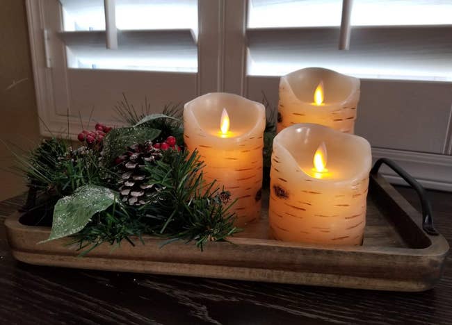 Reviewer image of three birch-patterned flameless candles with wavy tops turned on and sitting inside tray next to pinecones, leaves, and berries
