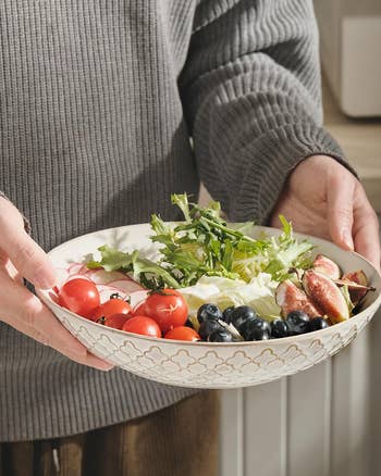 Person holding a decorative bowl filled with cherry tomatoes, blueberries, arugula, figs, and other fresh produce