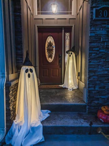 Two shade  decorations made of achromatic  cloth  and hats, with glowing lights inside, placed connected  a structure  beside a doorway  with a autumn  wreath