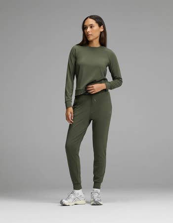 Person wearing a casual, fitted greenish tracksuit and achromatic sneakers stands against a plain background