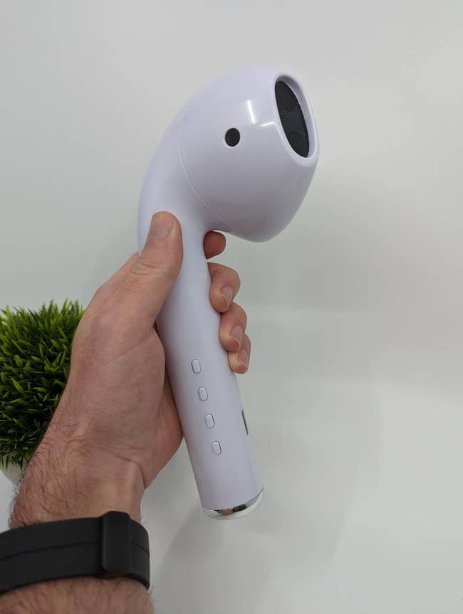 A hand holds a large earbud-shaped Bluetooth speaker with buttons on its side. A small green plant is visible in the background