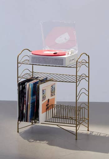 A gold wire vintage style storage rack with a record player on top and vinyls on the bottom shelf 