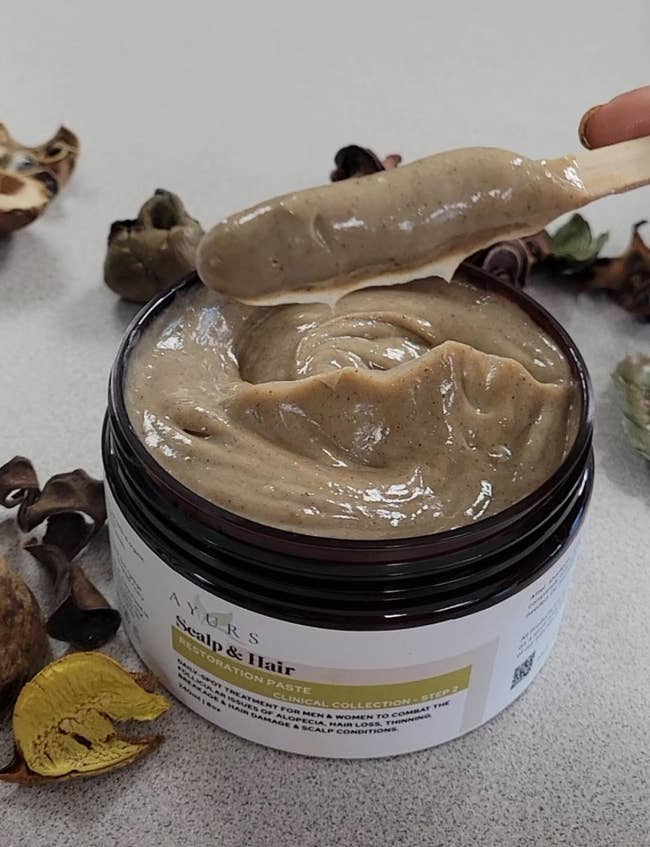 Open jar of scalp and hairsbreadth  restoration paste, creamy texture. Surrounded by dried botanicals, suggesting earthy  ingredients