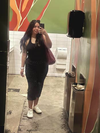 Person taking a reflector  selfie successful  a restroom, wearing a achromatic  sleeveless top, a polka-dot skirt, and achromatic  sneakers, carrying a ample  bag