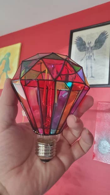 Stained glass-style airy bulb held successful a hand, surrounded by eclectic artwork connected a reddish wall