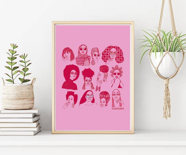 Graphic poster featuring diverse female faces illustration on a pink background, hanging on a wall beside a plant and stacked books