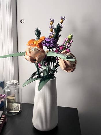 A vase holds an statement of LEGO flowers, including roses and lavender, connected a array with a jar of wealth successful the background