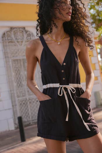 Person in a sleeveless romper with button details and a white tie belt, standing outdoors