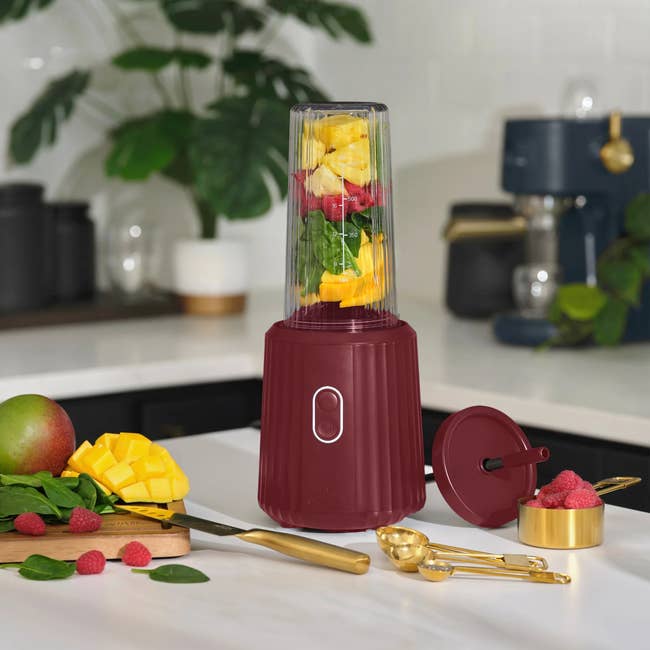 A room  countertop with a reddish  portable blender filled with fruit. Surrounding are mango, raspberries, spinach, and measuring spoons