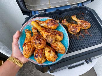 Grilled chicken drumsticks on a plate, next to more chicken on an open grill, suggesting a barbecue setting