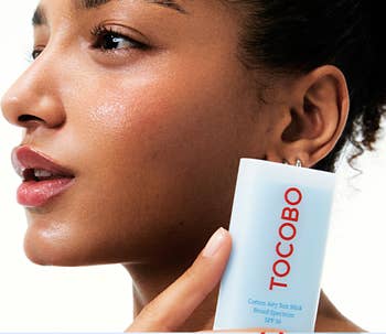 Person holding a Tocobo Cotton Airy Sun Stick, elaborate  with SPF 50, adjacent  to their cheek, highlighting skincare application