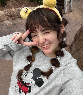 Person winking and making a bid    sign, wearing a Mickey Mouse sweatshirt and headband with ears, smiling outdoors
