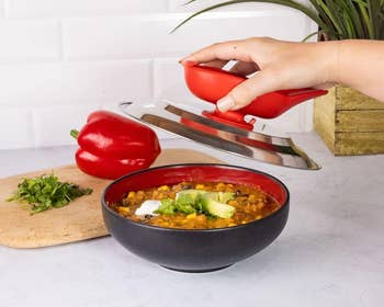 A hand lifts a red and silver magnetic lid off a bowl of soup with toppings. A chopped green herb and a red bell pepper are on a cutting board nearby