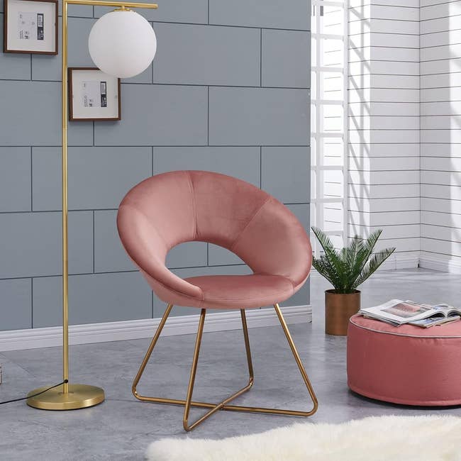 A modern surviving abstraction featuring a pinkish velvet seat with golden legs, a matching ottoman with magazines, a golden level lamp, framed photos, and a plant